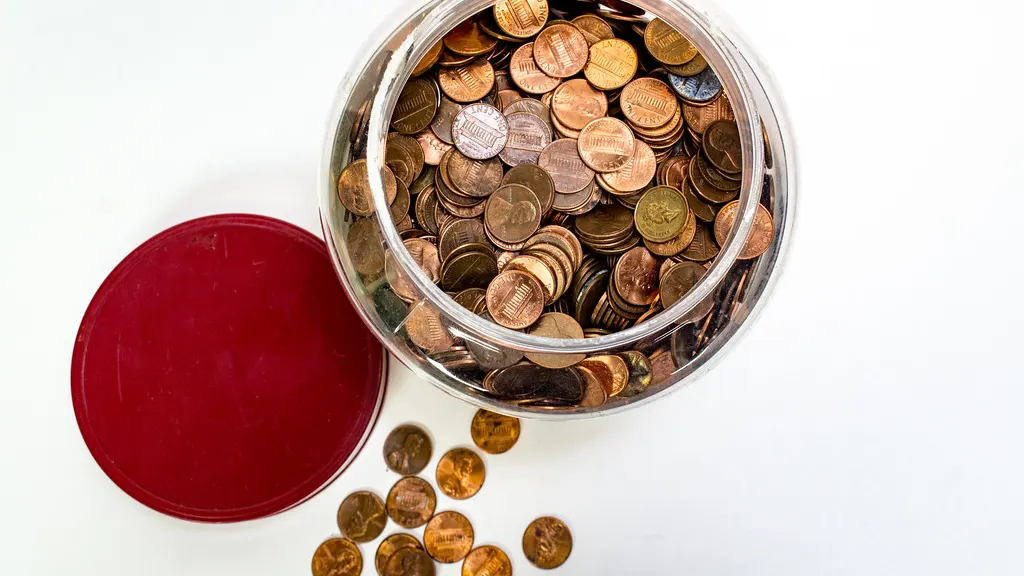 An open jar full of coins with its red lid beside it and pennies spilled on a white table