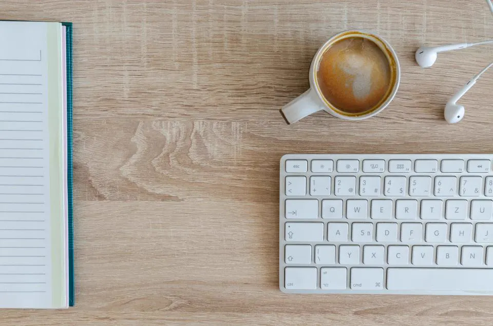 A cup of coffee, earbuds, a notebook and a keyboard on a wooden desk