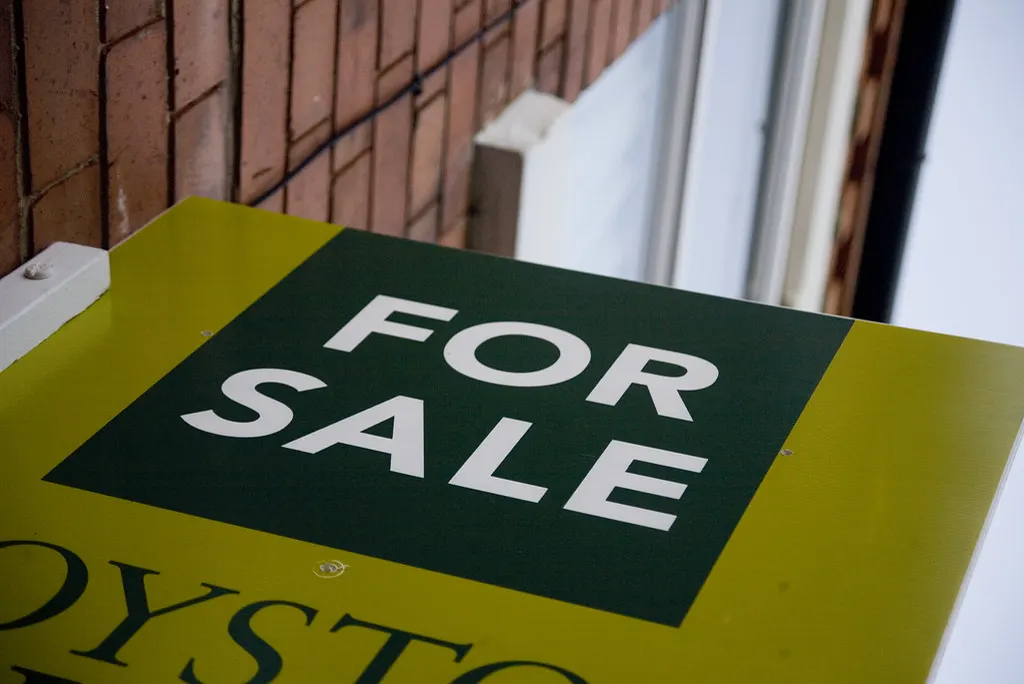 A green and yellow “For sale” sign in front of a brick house