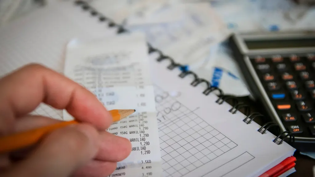 A hand holding a pencil over a till receipt, beside a notebook and a calculator