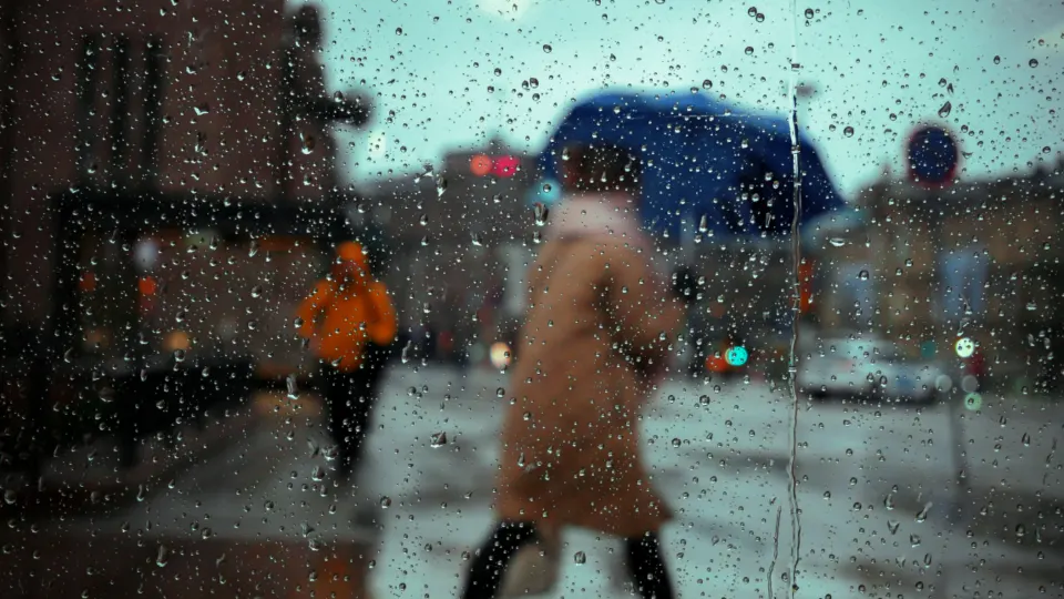 A person with a blue umbrella crossing a street, seen through a rain-streaked window